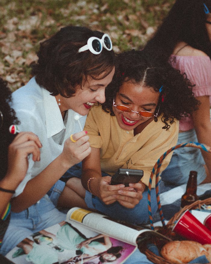our-story A group of happy young women enjoying a sunny picnic outdoors, engaging with a smartphone and laughing.